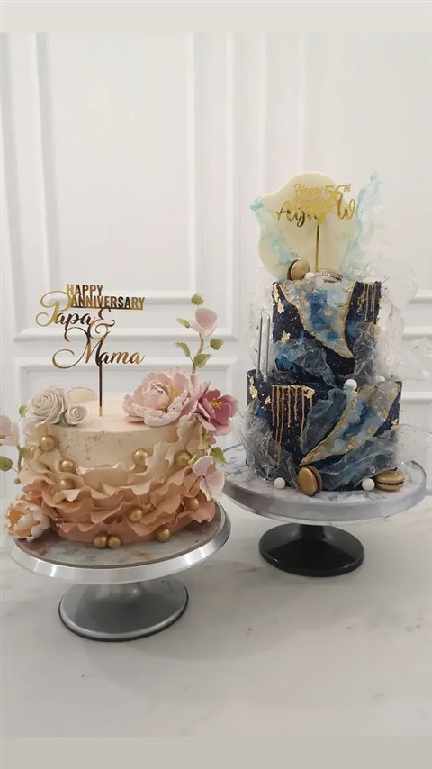 Kue anniversary blush dengan ruffle, bunga dan topper Happy Anniversary Papa & Mama, di samping kue biru geometris
