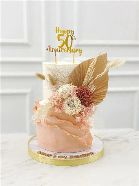 Kue dua tingkat ivory dan peach dengan bunga, daun palem kering dan topper Happy 50th Anniversary