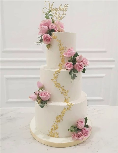 Kue pernikahan tiga tingkat ivory dengan mawar pink, daun hijau dan aksen emas