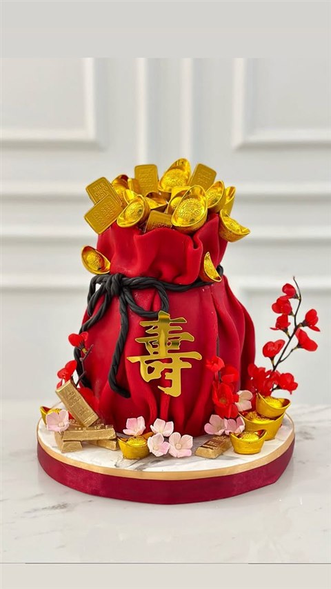 Kue berbentuk kantong merah dengan karakter panjang umur emas, hiasan batangan dan ingot emas, serta bunga