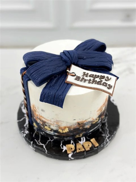 Kue krem dengan pita navy besar, aksen emas hitam, dan tulisan Happy Birthday