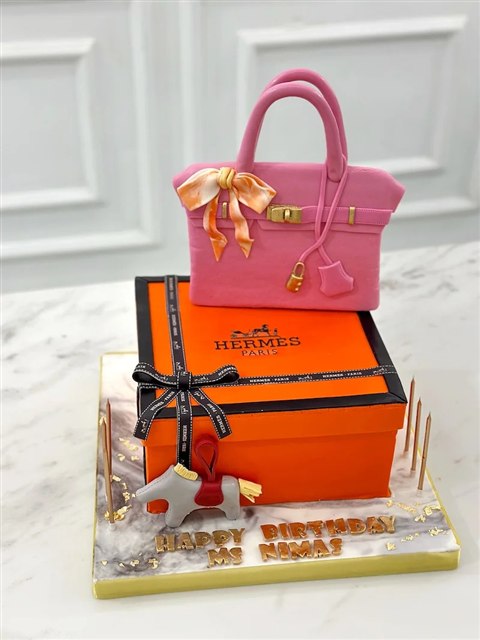 Kue berbentuk tas merah muda di atas kotak hadiah oranye dengan pita dan detail emas