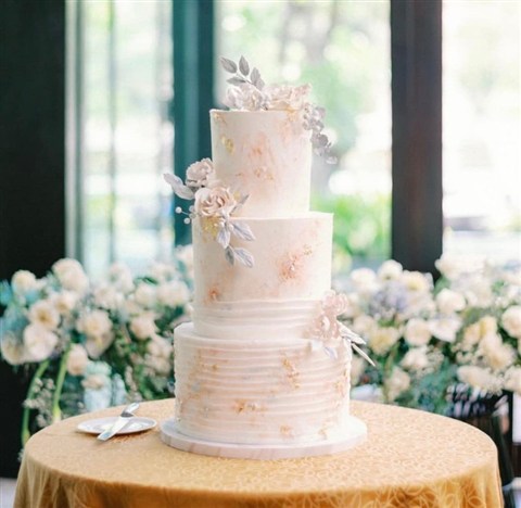 Kue wedding tiga tingkat berwarna putih dan blush di meja dengan rangkaian bunga