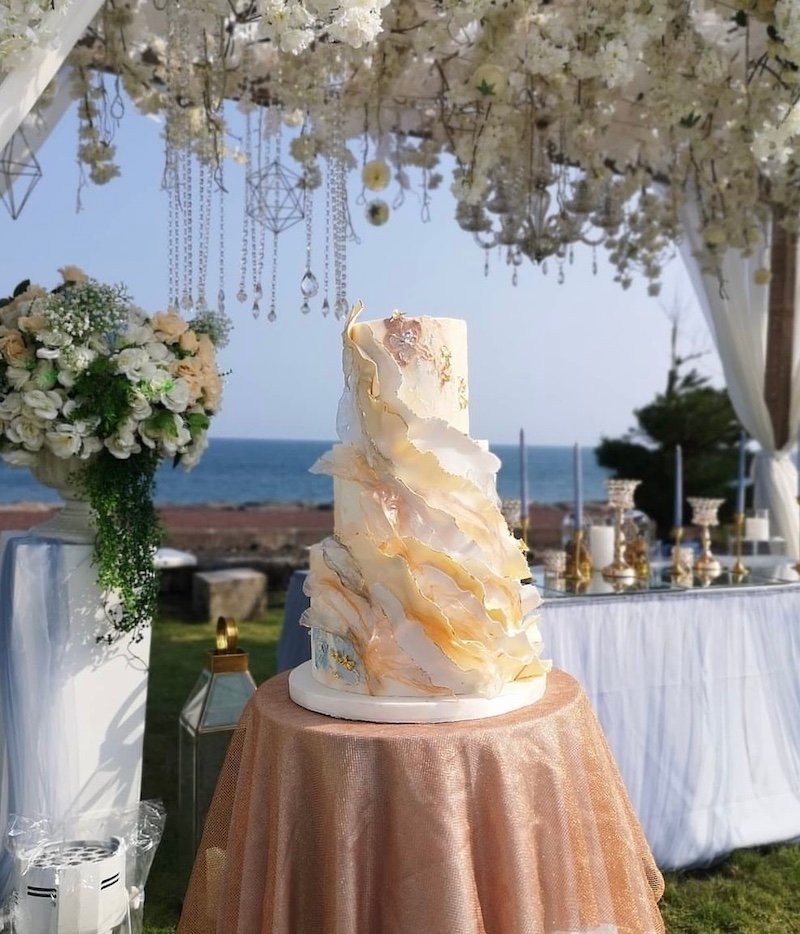 Kue wedding ivory berlapis kelopak dengan aksen emas di meja pernikahan berlatar laut
