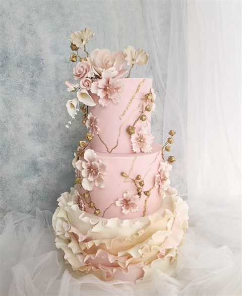 Kue pernikahan blush pink dengan bunga, garis emas, dan ruffle ivory